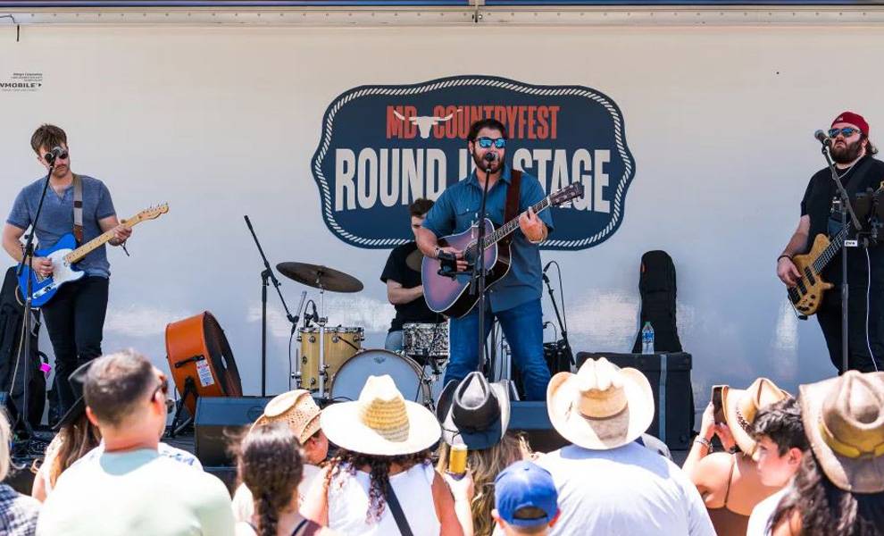 Una banda de música country actúa en vivo en un escenario al aire libre rotulado como «Round Up Stage», con músicos tocando guitarras y batería.