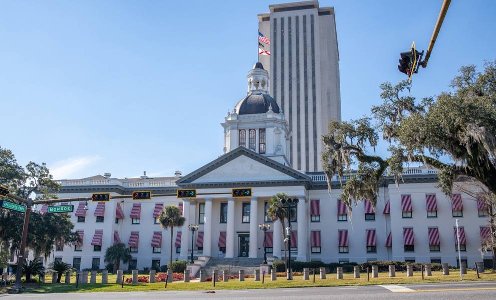 The Florida State Capitol building in Tallahassee, featuring the historic white domed structure in front of the modern high-rise capitol tower, with American and Florida flags flying above and a traffic light visible at the intersection.