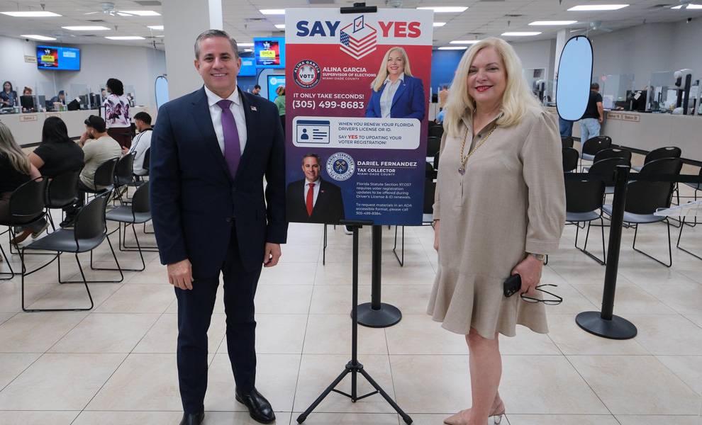 Un hombre con un traje azul marino y una mujer con un vestido beige permanecen de pie en el interior de una ajetreada oficina de servicios gubernamentales, junto a un letrero que dice «Di sí» y que contiene información sobre el registro de votantes. Detrás de ellos, varias personas se sientan en filas de sillas y esperan en los mostradores de atención bajo una brillante iluminación fluorescente.