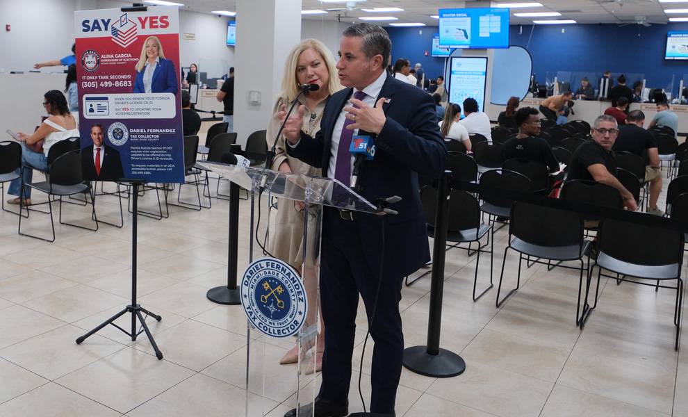 A man in a suit speaks at a podium with a microphone inside a crowded public service office while a woman stands behind him. A sign promoting voter registration is displayed nearby, and people are seated in waiting areas and being assisted at service desks in the background.