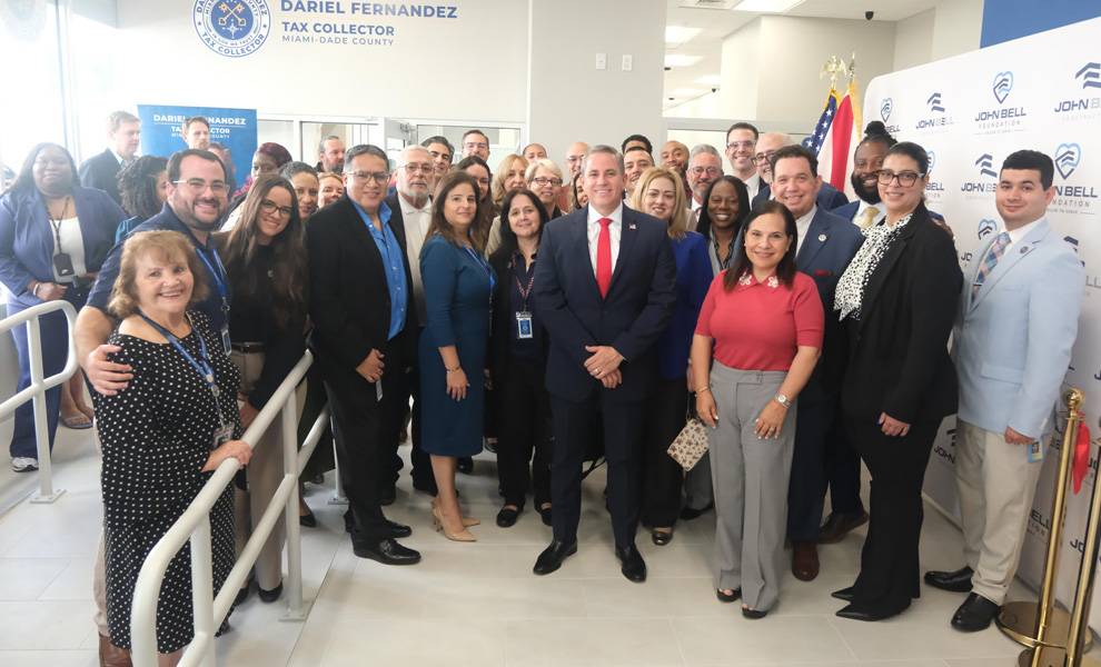Un grupo numeroso de empleados y líderes comunitarios posa para una foto dentro de una oficina gubernamental. Un hombre con traje azul marino y corbata roja se encuentra en el centro, rodeado de colegas. Un cartel en la pared dice "Dariel Fernández, Recaudador de Impuestos, Condado de Miami-Dade", y al fondo se ven un telón de fondo con la marca y una bandera estadounidense.