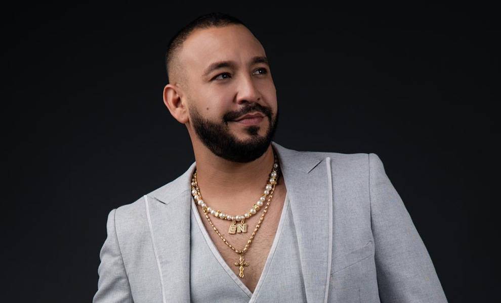 A man with a neatly trimmed beard poses against a dark background, wearing a light gray suit with a deep V-neck shirt.