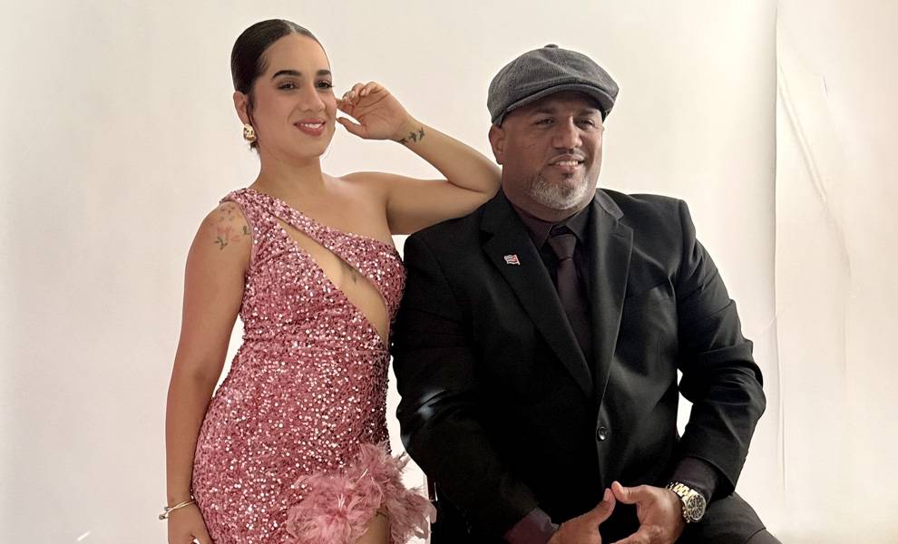 Una mujer con un vestido rosa de lentejuelas, de un solo hombro y con un detalle de plumas, está sentada junto a un hombre vestido con un traje negro y una gorra plana.