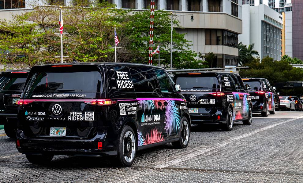 A line of black vans with colorful “Free Rides” and “Miami DDA Freebee” branding drive along a downtown street, with buildings, trees, and traffic in the background.