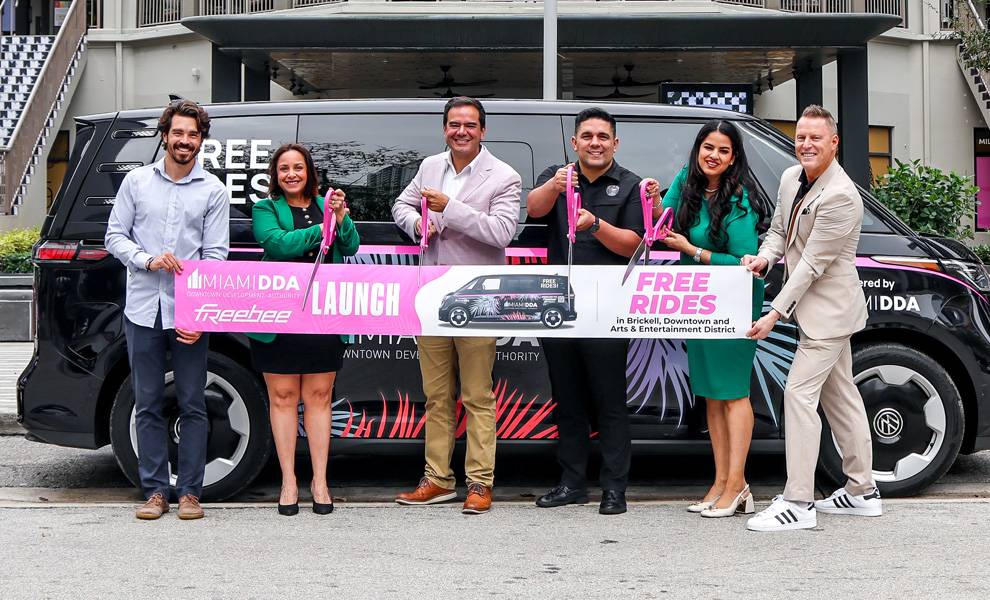 Un grupo de seis personas posa frente a una furgoneta rotulada con la marca "Free Rides", sonriendo y sosteniendo una gran cinta ceremonial y unas tijeras durante un evento de lanzamiento.