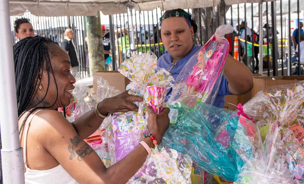 Dos mujeres permanecen de pie bajo una carpa en un evento comunitario para The Caring Place, clasificando e intercambiando coloridas cestas de regalo envueltas en celofán transparente.