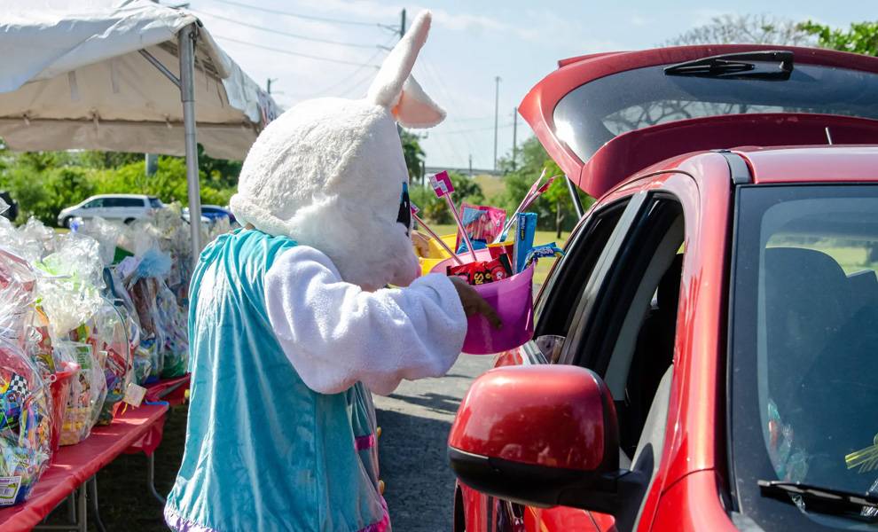 A person dressed in a white Easter bunny costume hands a pink basket filled with toys and treats through the window of a red car at a drive-through giveaway at Giving Thanks on Good Friday at The Caring Place.