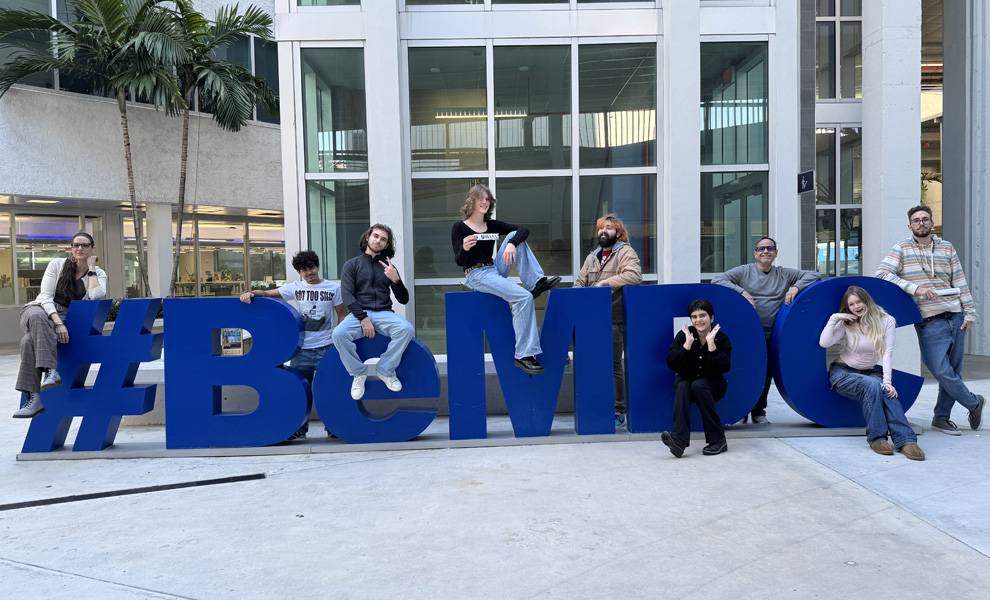Un grupo de estudiantes posa al aire libre frente a grandes letras azules que forman la palabra “#BeMDC” en un campus universitario.