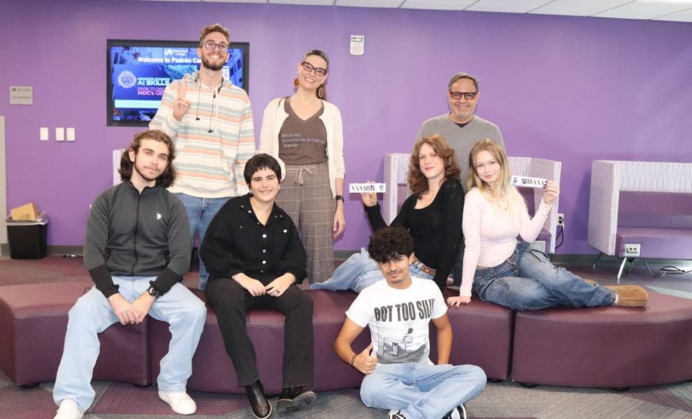 A group of students and a faculty member pose together inside a campus lounge with purple walls with an Urbana sticker.