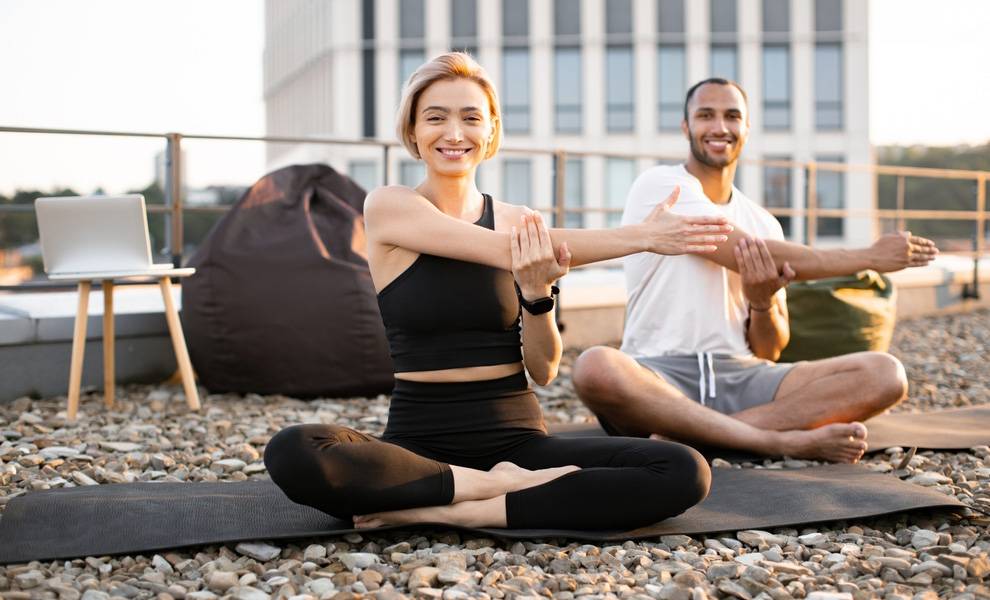 Two adults sit cross-legged on yoga mats on a rooftop at sunset, smiling while stretching one arm across their chest.