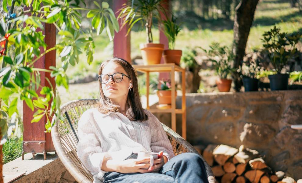 Mental health in the workplace: How Aetna™ is supporting Miami-Dade County employees beyond traditional coverage 1 A woman wearing glasses relaxes in a wicker chair on a sunlit porch surrounded by green plants. She holds a cup in her hands with her eyes closed, appearing calm and peaceful, with potted plants and stacked firewood nearby prioritizing her mental health.