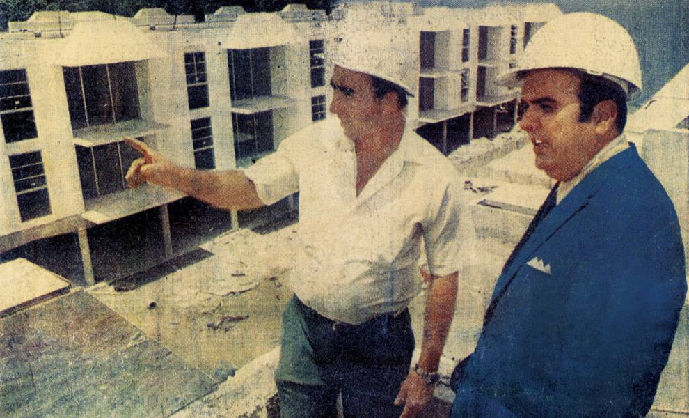 Two men wearing white construction hard hats stand at a building site, looking over a partially completed multi-story structure.