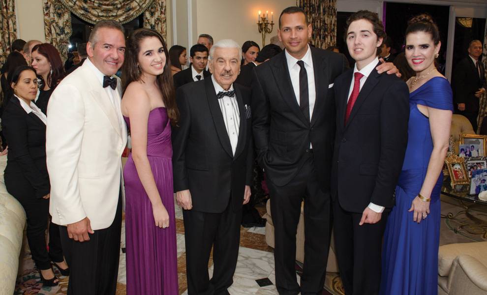 Jose Milton in a tuxedo stands at the center of a group at an indoor formal gathering, surrounded by well-dressed family members and guests in evening attire, smiling for a photo.