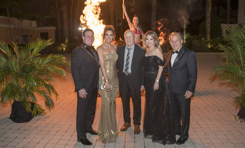 Five formally dressed adults pose outdoors at night on a paved courtyard, smiling in front of a large fire feature. A fire performer is visible in the background, and palm trees frame the scene, creating an elegant South Florida Hispanic Chamber gala atmosphere.