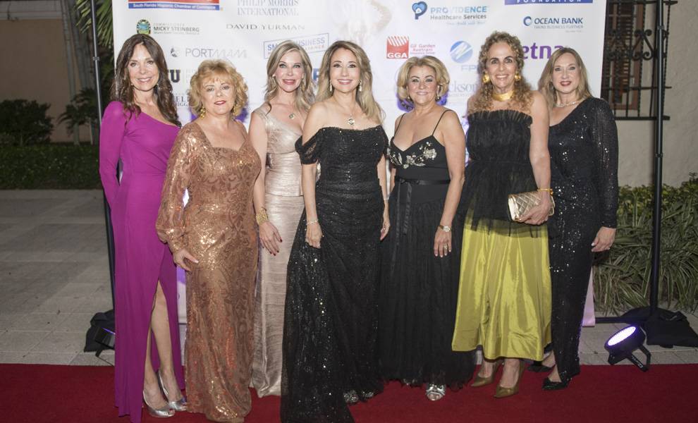 Siete mujeres posan juntas sobre una alfombra roja, frente a un telón de fondo con los logotipos de los patrocinadores, en una gala de la SFLHCC.