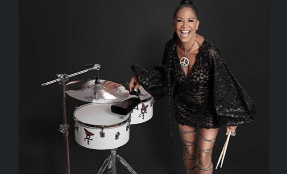Singer Sheila E in a black patterned mini dress with flowing sleeves stands beside a timbales setup, holding drumsticks in one hand.