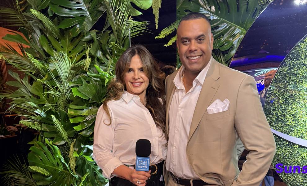 Rosi Rosell holding a “Calle Ocho News” microphone stands beside a man in a beige suit as they pose together at a Sunset Smile Creations event.