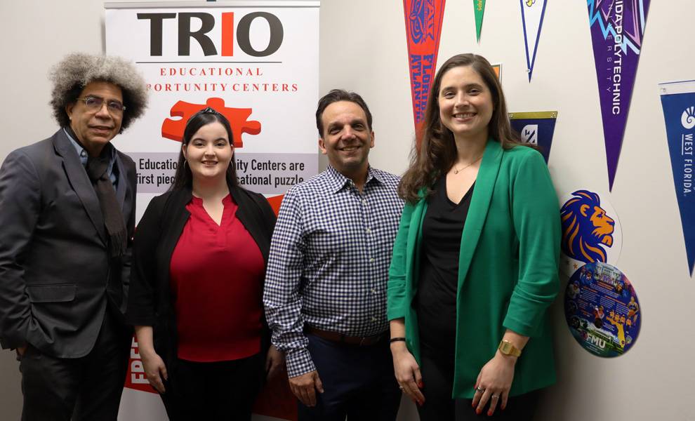 Cuatro profesionales posan junto a un banner de los Centros de Oportunidades Educativas TRIO. El banner destaca servicios gratuitos tales como asistencia con la ayuda financiera, ayuda con las admisiones universitarias, asesoramiento profesional e información sobre el GED. El grupo viste atuendo formal de negocios y sonríe en el entorno interior de una oficina.