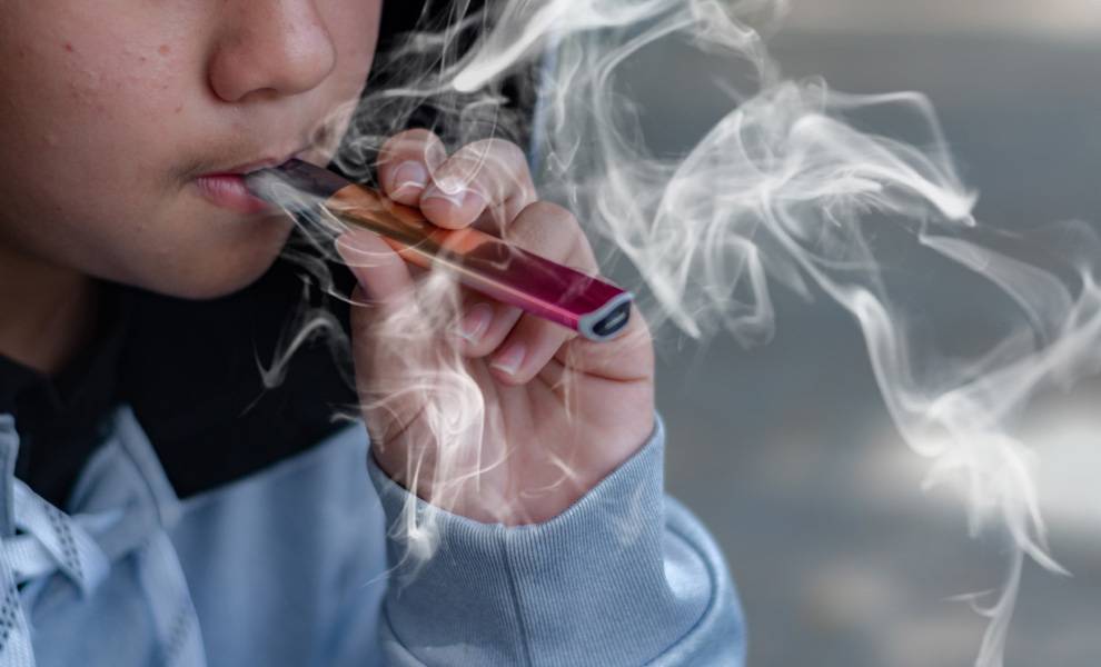 Florida schools shift approach as youth vaping concerns grow 1 Close-up of a young person holding a small vaping device to their mouth, exhaling a thick cloud of vapor that partially obscures their face.