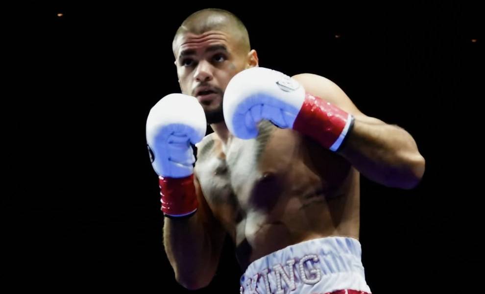 A focused male boxer with a shaved head and trimmed beard raises his gloves in a defensive stance inside a dimly lit ring.
