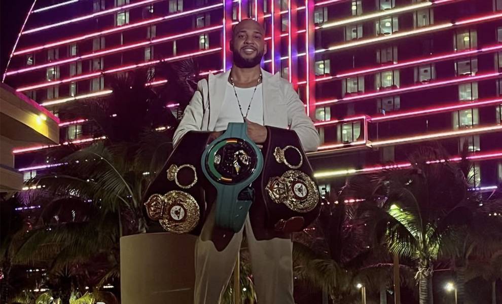 Un hombre sonriente se encuentra al aire libre, de noche, frente a un edificio brillantemente iluminado con luces de neón rosas y rojas, sosteniendo múltiples cinturones de campeonato de boxeo que lleva colgados sobre los brazos y los hombros.