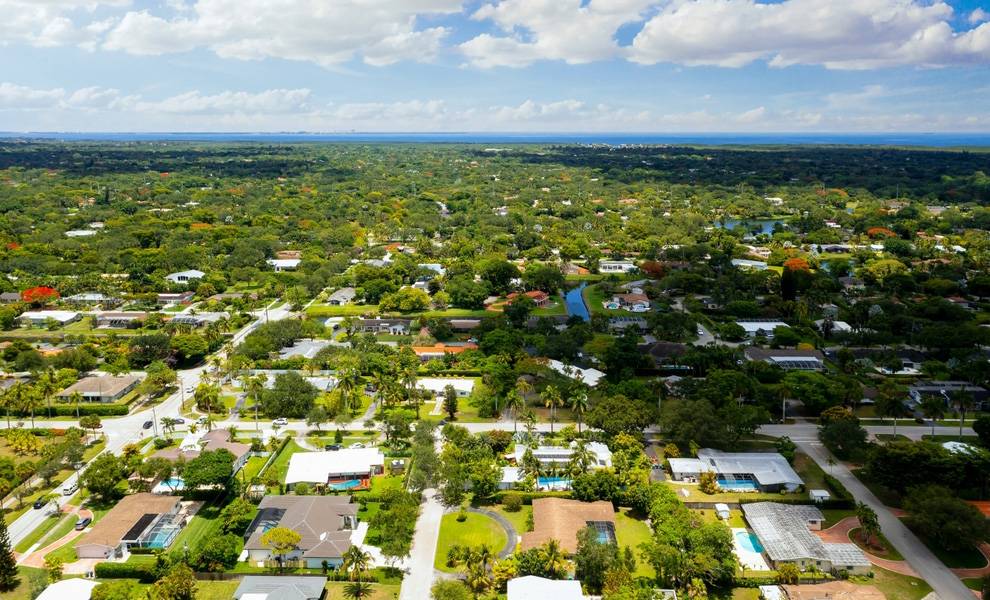 Vista aérea de un exuberante vecindario suburbano de Miami con casas unifamiliares, calles arboladas y piscinas en el patio trasero que se extienden hacia una costa lejana bajo un cielo parcialmente nublado.