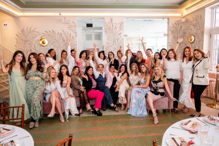 A large group of women pose together inside an elegant, softly lit venue, many seated and others standing behind them with hands raised in celebration.