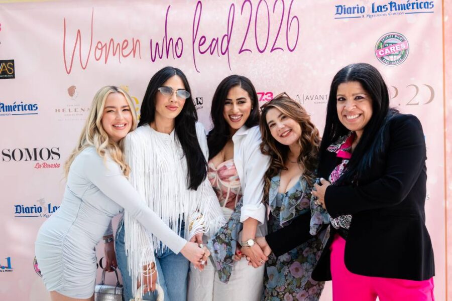 Five women stand closely together in front of a branded event backdrop, smiling and joining hands in a gesture of unity at the Women Who Lead 2026 event.