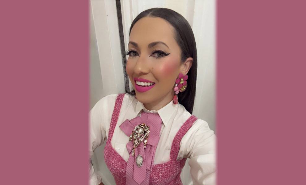 Close-up selfie of a smiling woman wearing a white blouse under a pink textured dress with a large decorative brooch and bow tie at the collar.