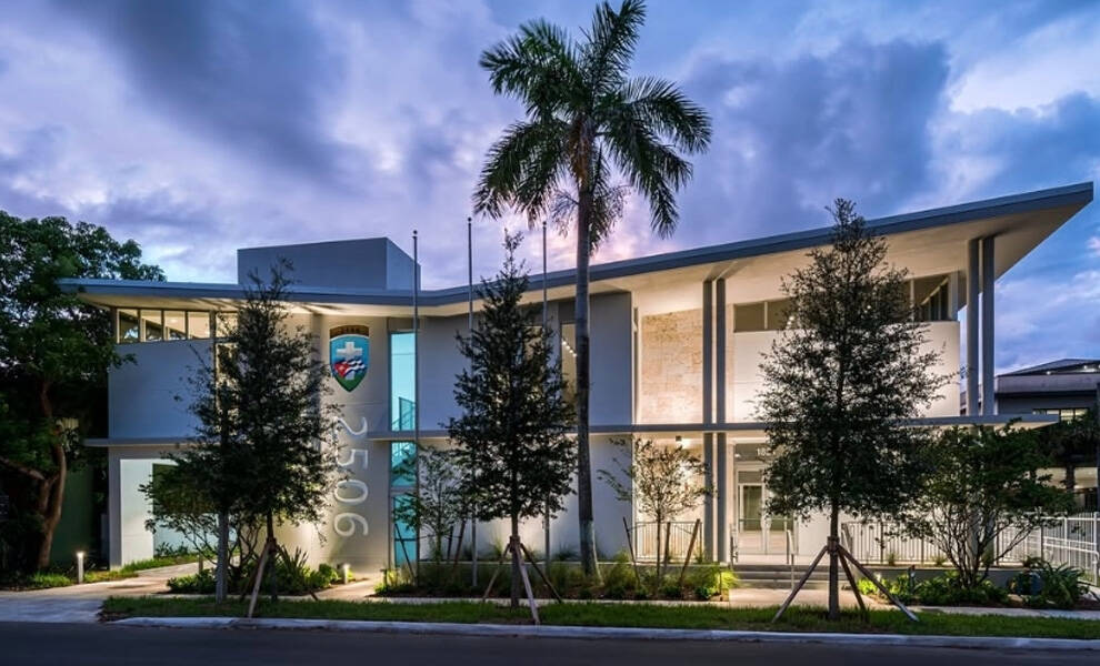 The Bay of Pigs Museum. A tall palm tree stands in front, along with neatly landscaped greenery and small trees. The building’s facade features a vertical address number and a colorful crest near the entrance.