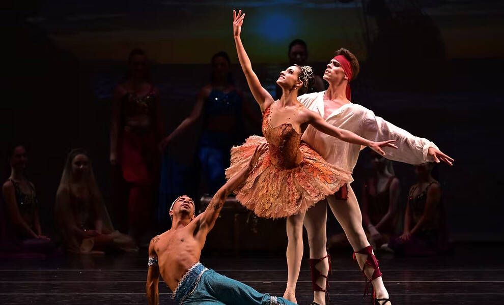 CCBM Ballet dancers perform Le Corsaire Suite on stage under dramatic lighting, with a central ballerina in a detailed orange tutu extending her arm gracefully while two male dancers pose alongside her.