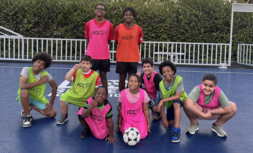 A group of children and two older teens pose together on an outdoor sports court, smiling and wearing colorful training pinnies with “FCC” on the front. A soccer ball rests in front as they kneel and stand in a team formation, with greenery and a fence in the background.