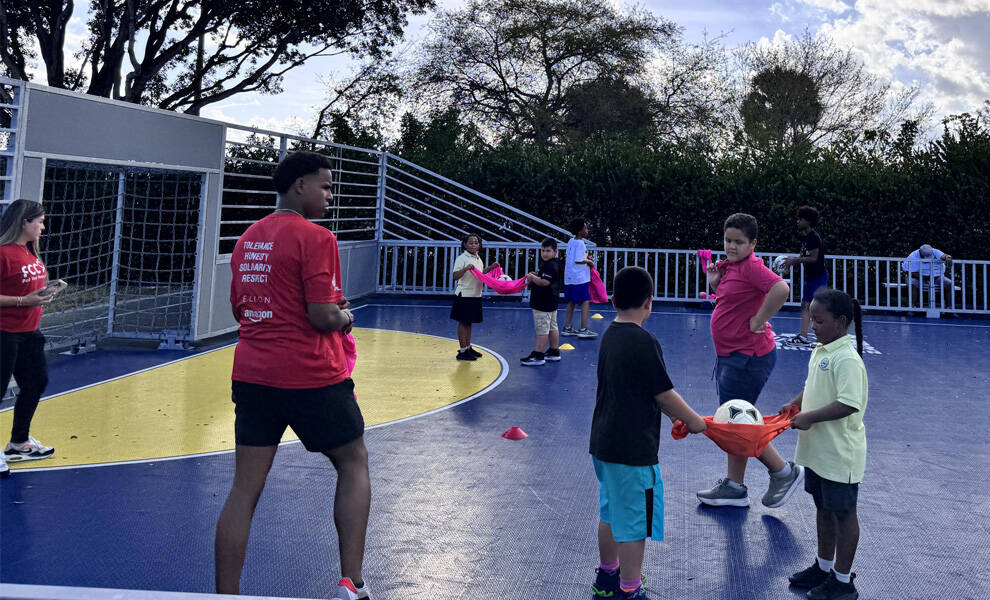 Children participate in a group activity on an outdoor court, using a red cloth to carry a soccer ball together while others watch and play nearby. A coach or older youth in a red shirt stands guiding the activity, with cones and a goal visible in the background.