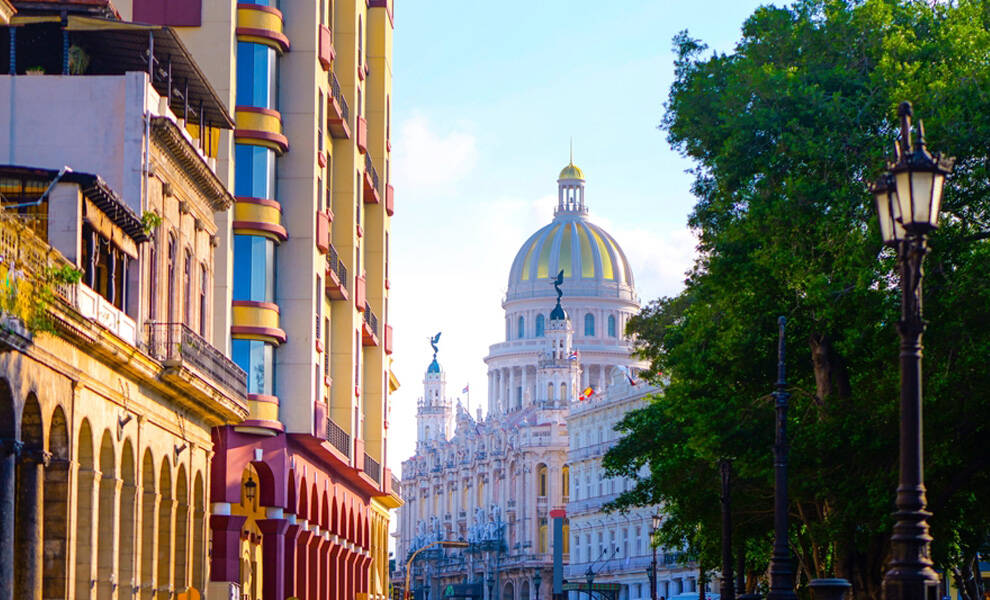 Colorful colonial buildings line a sunlit Cuban street, leading the eye toward a grand domed capitol building in the distance. Lush green trees and vintage street lamps frame the scene, capturing a vibrant urban view with historic architecture.