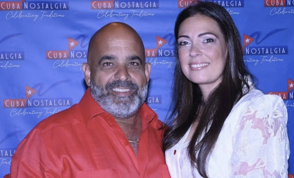 A man in a red shirt and a woman in a light-colored dress smile for a close-up photo in front of a “Cuba Nostalgia” step-and-repeat backdrop.