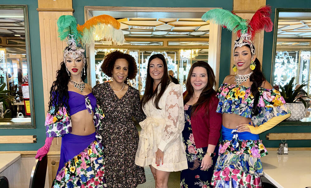 Two showgirls in vibrant, floral costumes with feathered headpieces stand on either side of three women in dresses, all posing together inside a brightly decorated Cuba Nostalgia event with mirrors and warm lighting.