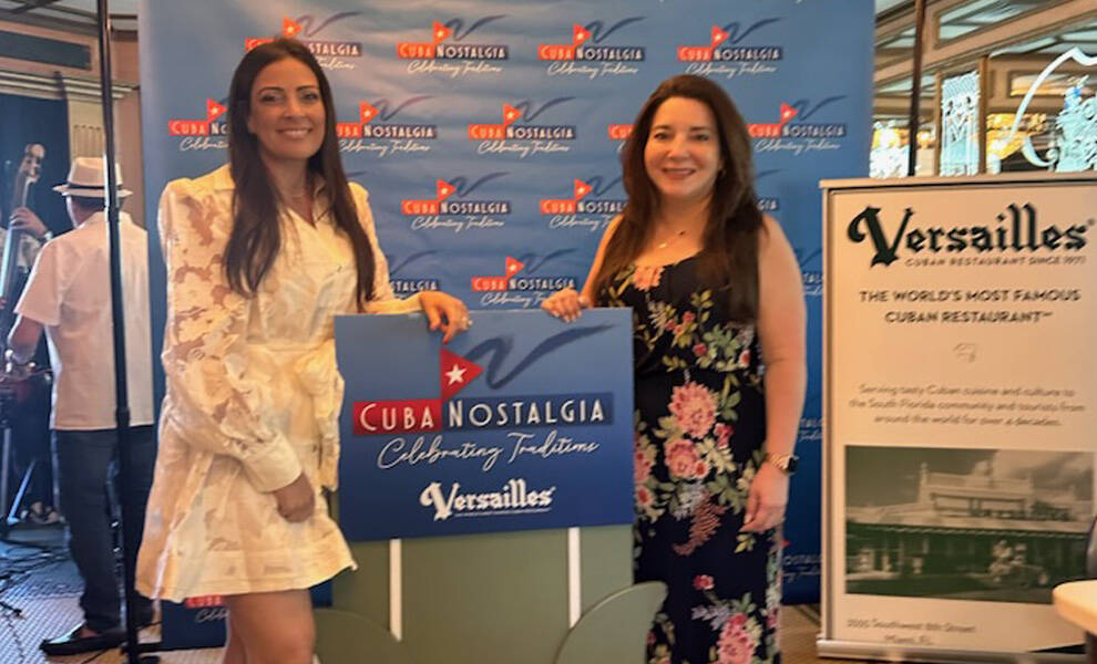 Dos mujeres posan frente a un telón de fondo de "Cuba Nostalgia" sosteniendo un letrero que dice "Versailles", con un panel informativo cercano que destaca el icónico restaurante cubano, mientras otros invitados y músicos aparecen al fondo.