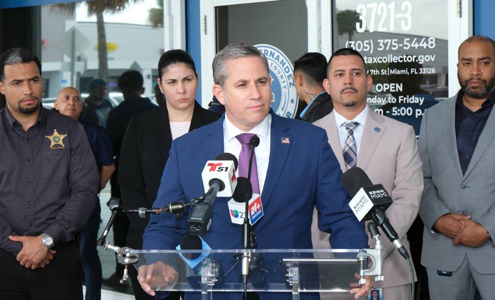 El Recaudador de Impuestos del condado de Miami-Dade, Dariel Fernandez, habla desde un podio con múltiples micrófonos frente a un edificio, flanqueado por funcionarios y personal.