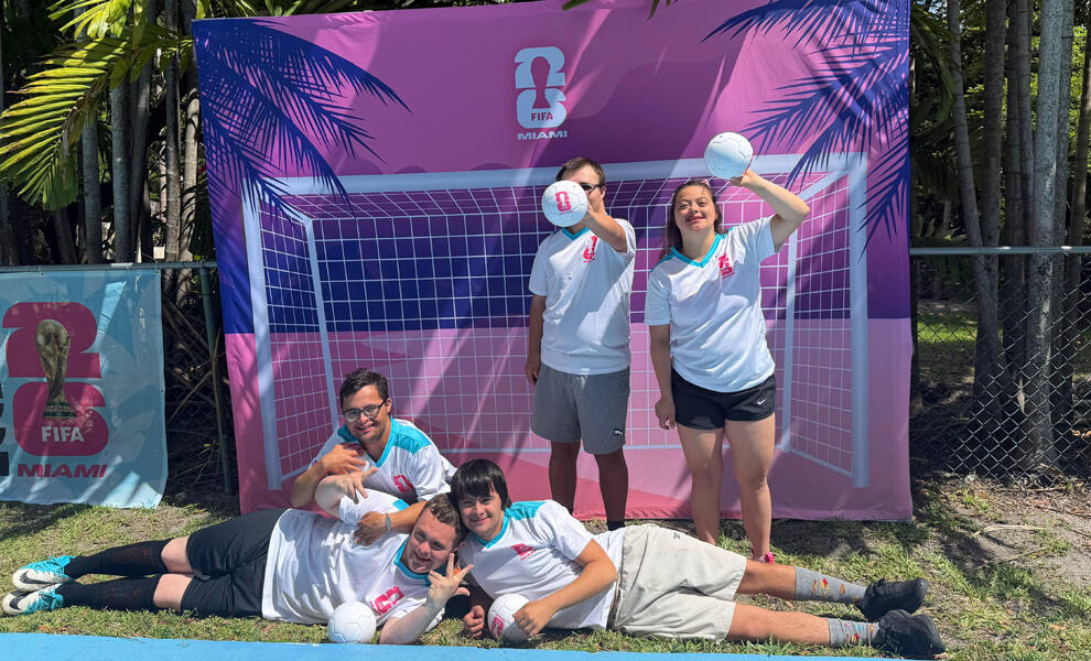 Un grupo de jóvenes participantes posa al aire libre frente a un telón de fondo rosa con temática de fútbol de la FIFA 2026™ Miami, sosteniendo balones de fútbol y sonriendo, mientras descansan y permanecen de pie sobre el césped, vistiendo ropa deportiva informal.