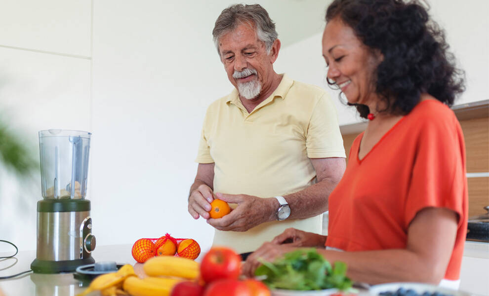 Un hombre y una mujer que llevan un estilo de vida saludable se encuentran en una cocina luminosa, preparando fruta fresca juntos, con una licuadora sobre la encimera e ingredientes como plátanos, manzanas y naranjas dispuestos a la vista.