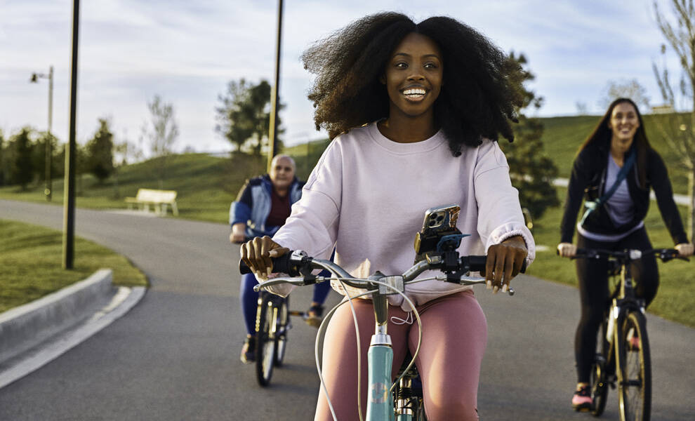 Tres personas pasean en bicicleta por un sendero pavimentado de un parque en un día soleado, con una mujer sonriente en primer plano y otras dos siguiéndola por una ruta cubierta de hierba y bordeada de árboles.
