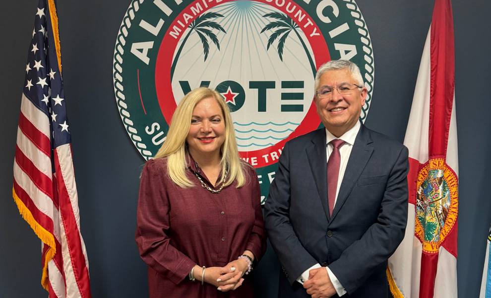 Miami-Dade Supervisor of Elections Alina Garcia and Peru’s Consulate stand smiling in front of a Miami-Dade County “I Voted” backdrop, with the U.S. flag on the left and the Florida state flag on the right.