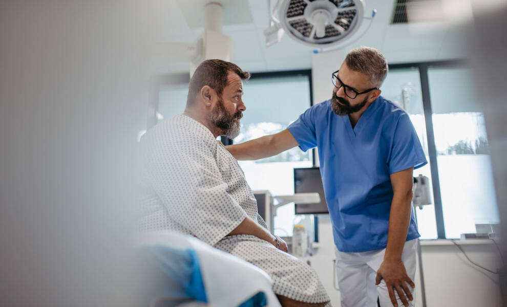 Un profesional de la salud, vestido con uniforme médico azul, conversa con suavidad con un paciente sentado que lleva una bata de hospital, en una sala de exploración clínica. El paciente se muestra atento mientras el profesional se inclina hacia él con actitud de apoyo; al fondo, se aprecian equipos médicos y la iluminación del techo.