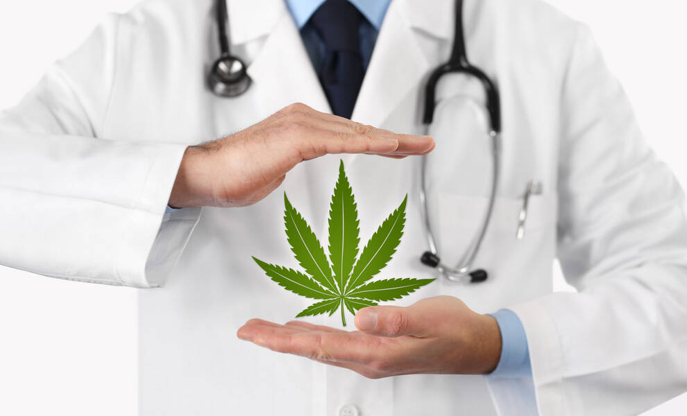 A doctor wearing a white coat and stethoscope holds their hands around a medical marijuana leaf, symbolically framing it as part of medical care or treatment.