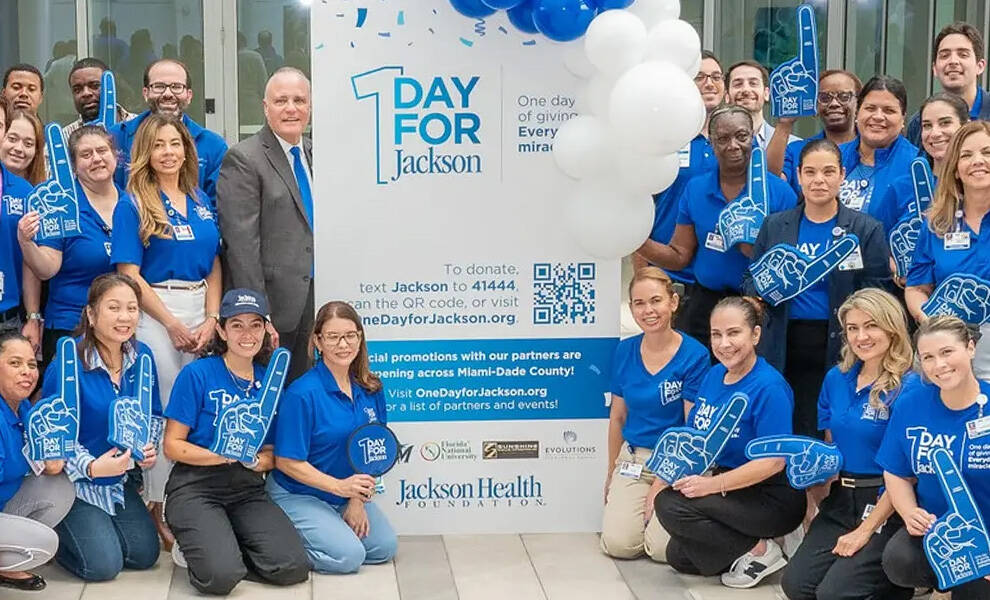 A large group of healthcare staff and supporters wearing blue shirts pose around a “1 Day for Jackson” sign and balloon display, many holding blue foam fingers, promoting a community fundraising campaign for Jackson Health Foundation.