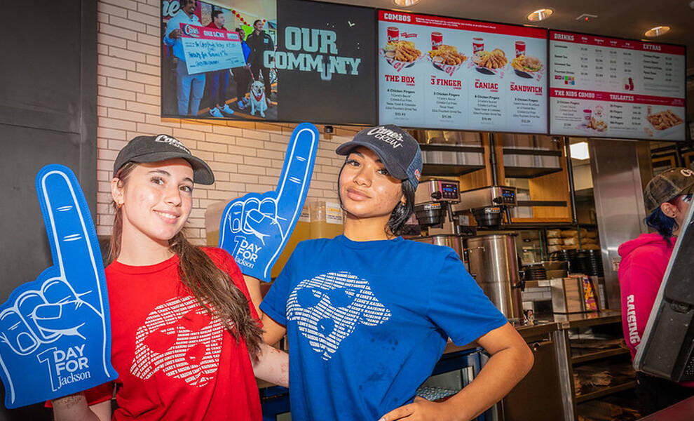 Two restaurant employees wearing caps and branded shirts stand inside a fast-food restaurant, smiling and holding blue “1 Day for Jackson” foam fingers, with menu boards and a counter visible behind them.