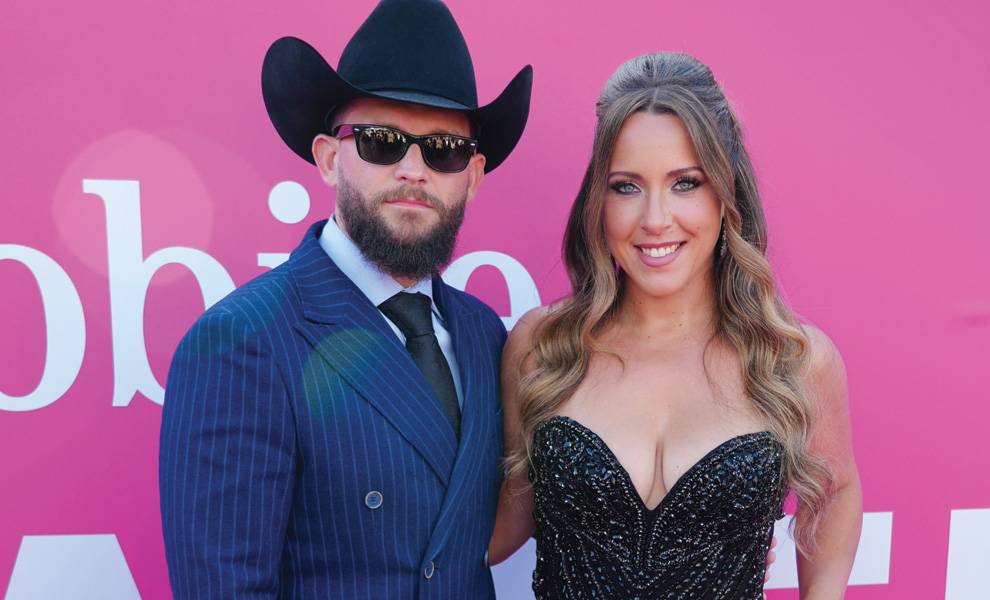 Un hombre con un sombrero de vaquero negro y gafas de sol se encuentra junto a una mujer con un vestido formal negro, frente a un telón de fondo rosa para eventos.