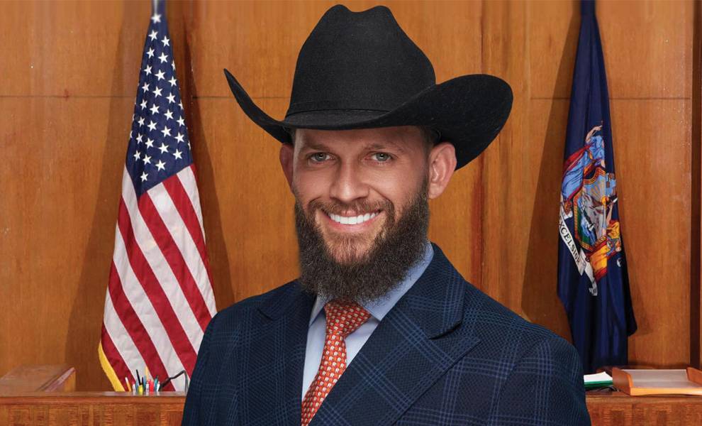 Home Miami (EN) New 55 Robert Kelbert wearing a black cowboy hat and suit smiles in a courtroom setting with U.S. and state flags behind him.