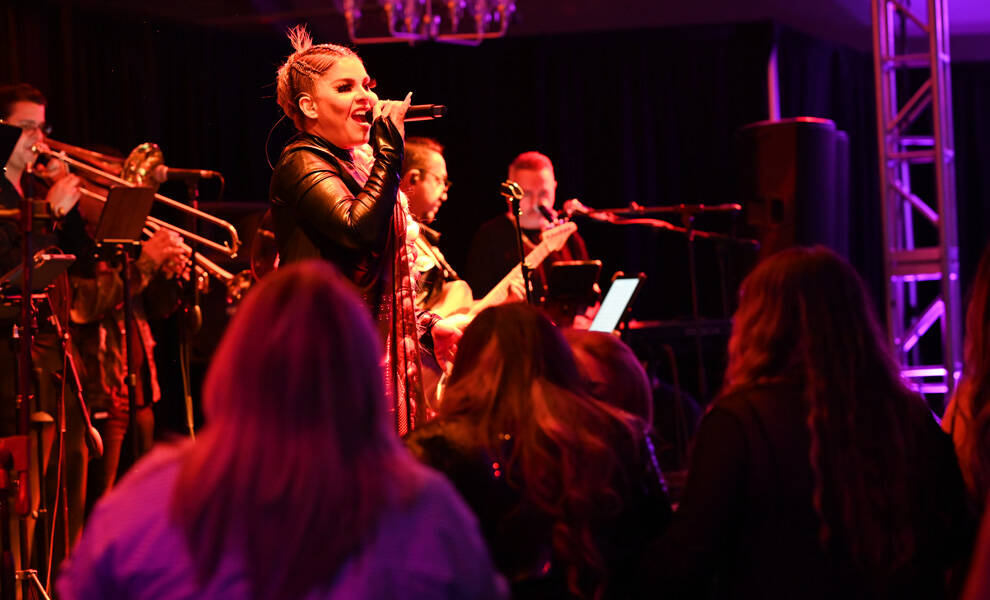 A female singer performs on stage at Seminole Classic Casino under red and purple lighting, holding a microphone and singing energetically while a live band with brass instruments plays behind her. Audience members are visible in the foreground, watching the performance.