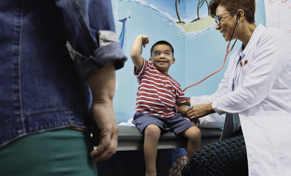 A smiling young boy sits on an exam table flexing his arm while a healthcare provider listens to his heartbeat with a stethoscope. An adult stands nearby, partially visible, as the brightly decorated pediatric room creates a friendly, reassuring environment.